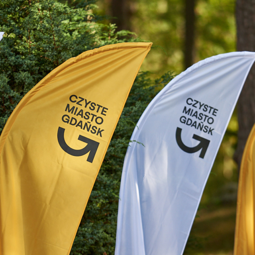 Flagi promujące inicjatywę "Czyste Miasto Gdańsk" w kolorach żółtym, białym i czarnym, umieszczone w lesie, symbolizujące dbałość o czystość i ekologię w Gdańsku.